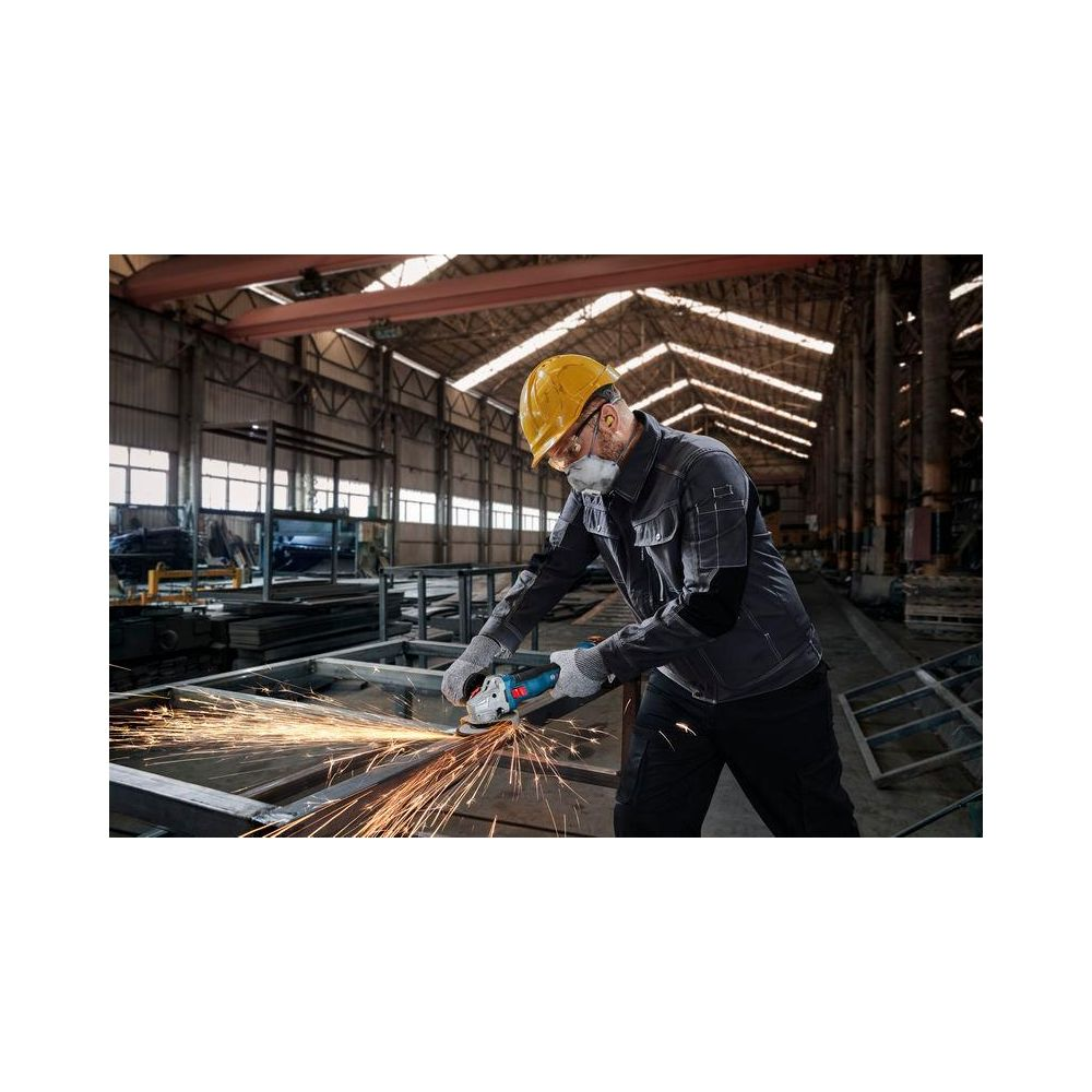 Mann mit gelbem Helm schleift Metall, Funken fliegen in Fabrik.