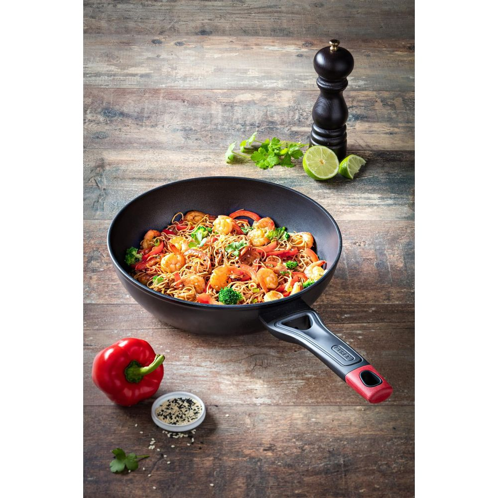 A wok filled with noodles, shrimp, and vegetables sits on a wooden table.