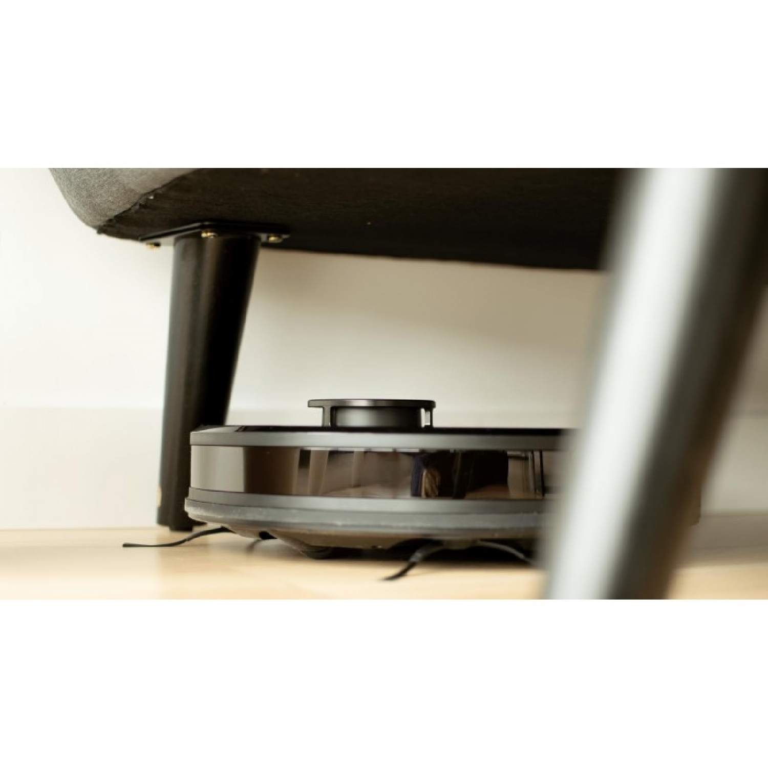 Un robot aspirapolvere nero pulisce sotto un mobile con gambe nere.