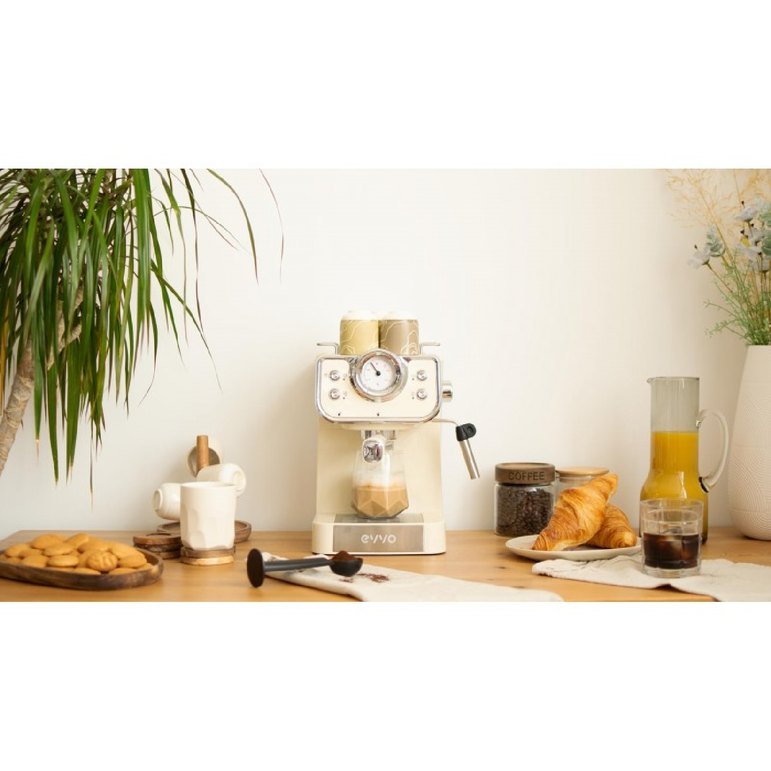 Una máquina de espresso color crema prepara café junto a cruasanes, galletas, zumo y un vaso de café helado.