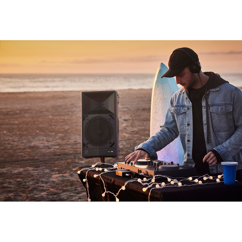 DJ mit Kopfhörern legt am Strand bei Sonnenuntergang auf. Lautsprecher, Surfbrett im Hintergrund.