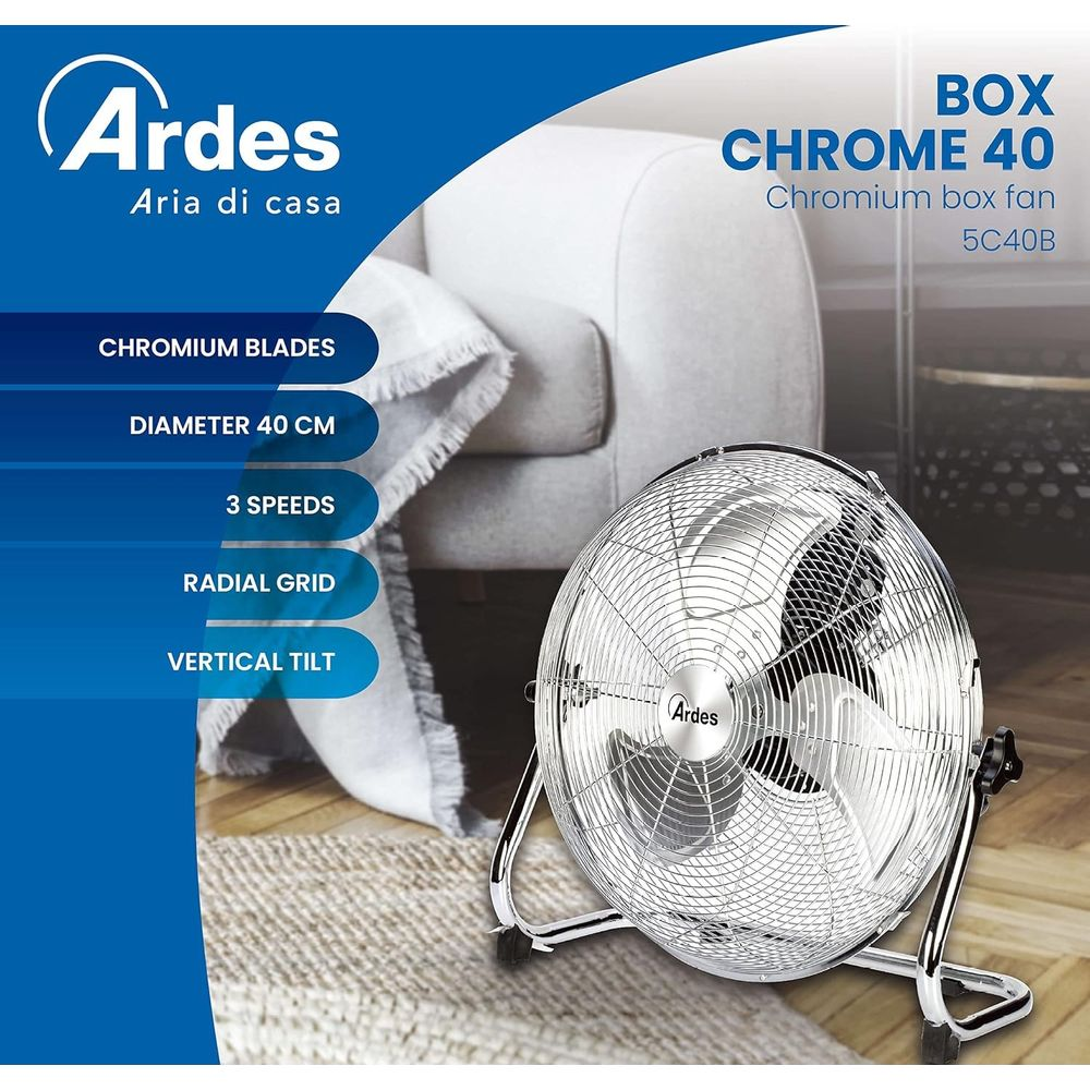 Een zilveren Ardes boxventilator met chromen bladen staat op een vloerkleed voor een fauteuil.