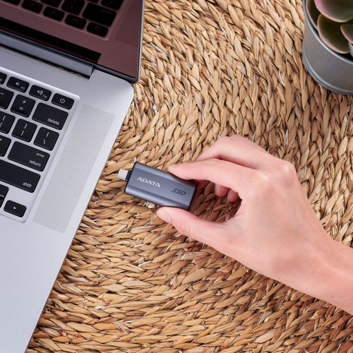 Una mano tiene un SSD ADATA grigio vicino a un laptop argentato su un tappetino intrecciato.