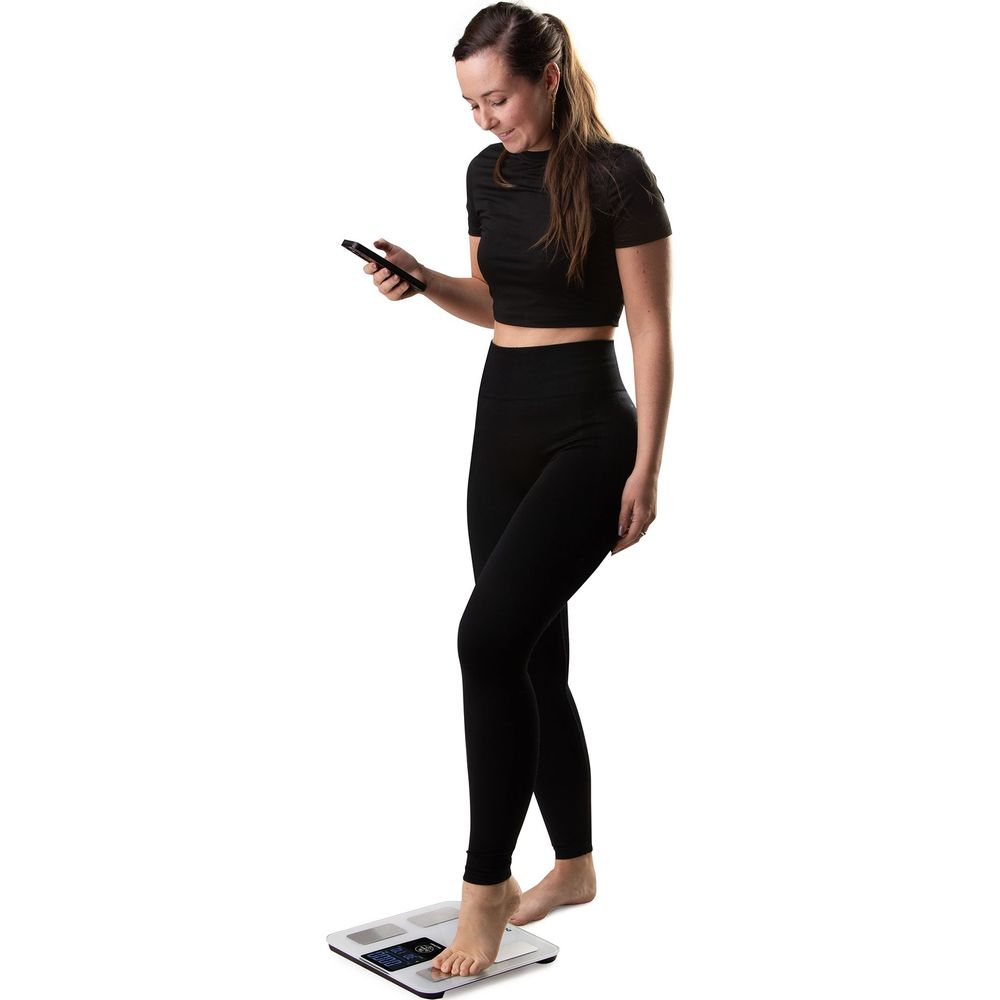 Una donna in nero è in piedi su una bilancia, guarda il suo telefono.