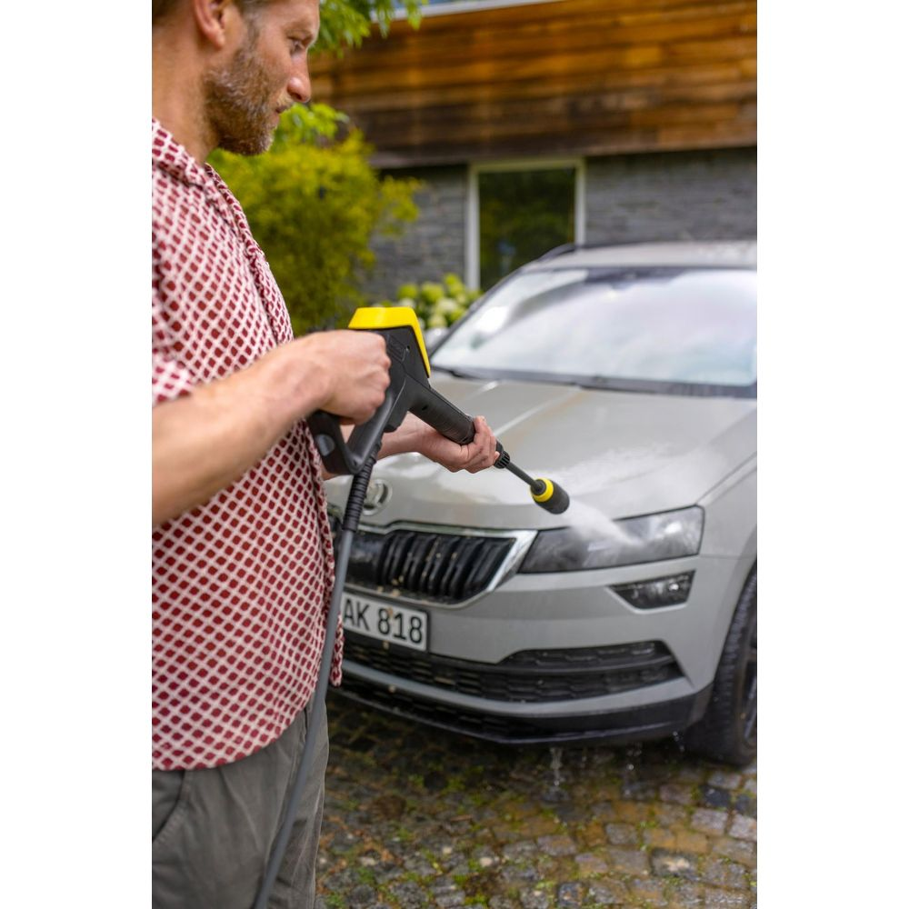 Mann reinigt Auto mit Hochdruckreiniger. Graues Auto, Haus im Hintergrund.