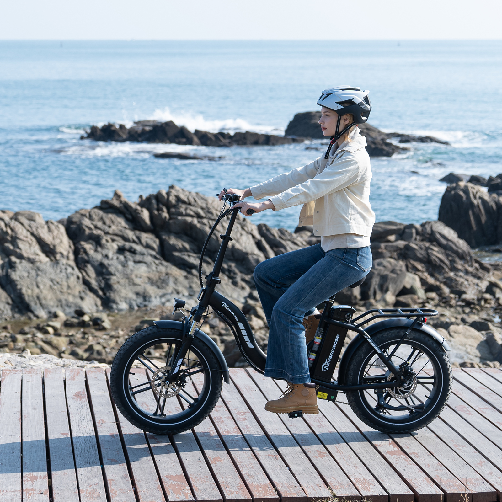 Eine Frau mit Helm fährt ein schwarzes E-Bike auf einem Holzweg am Meer.