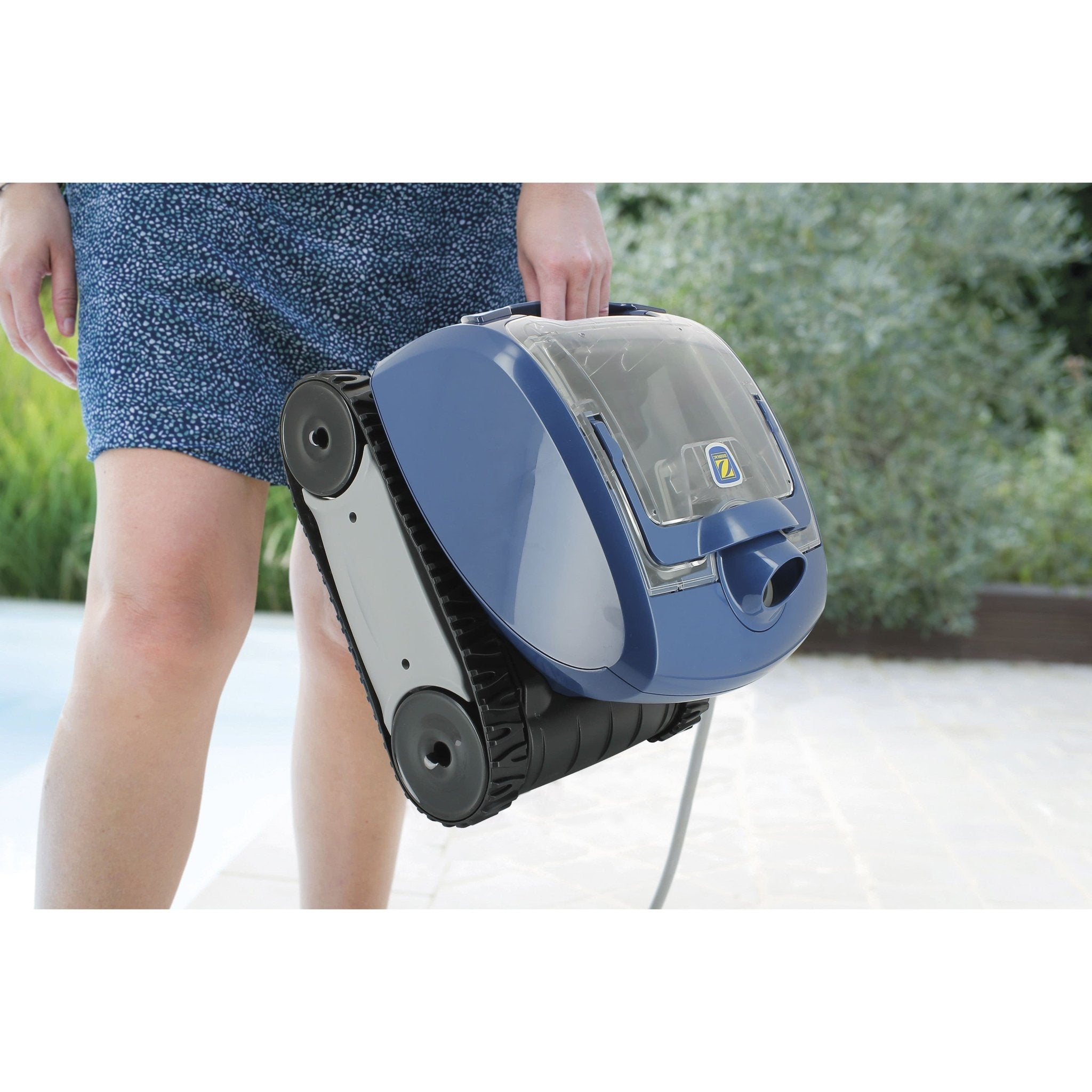 Una persona sujeta un robot limpiafondos azul con orugas de goma junto a una piscina.