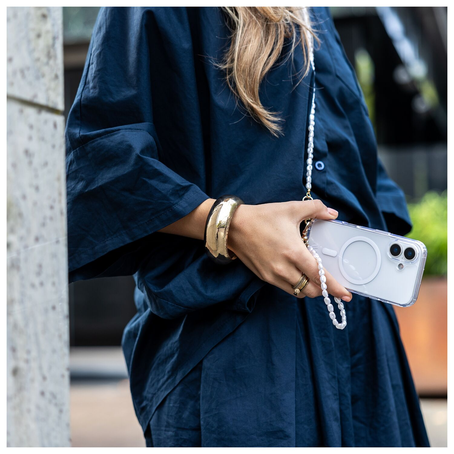 Een vrouw in een donkerblauwe jurk houdt een witte smartphone met parelketting vast en draagt gouden sieraden.