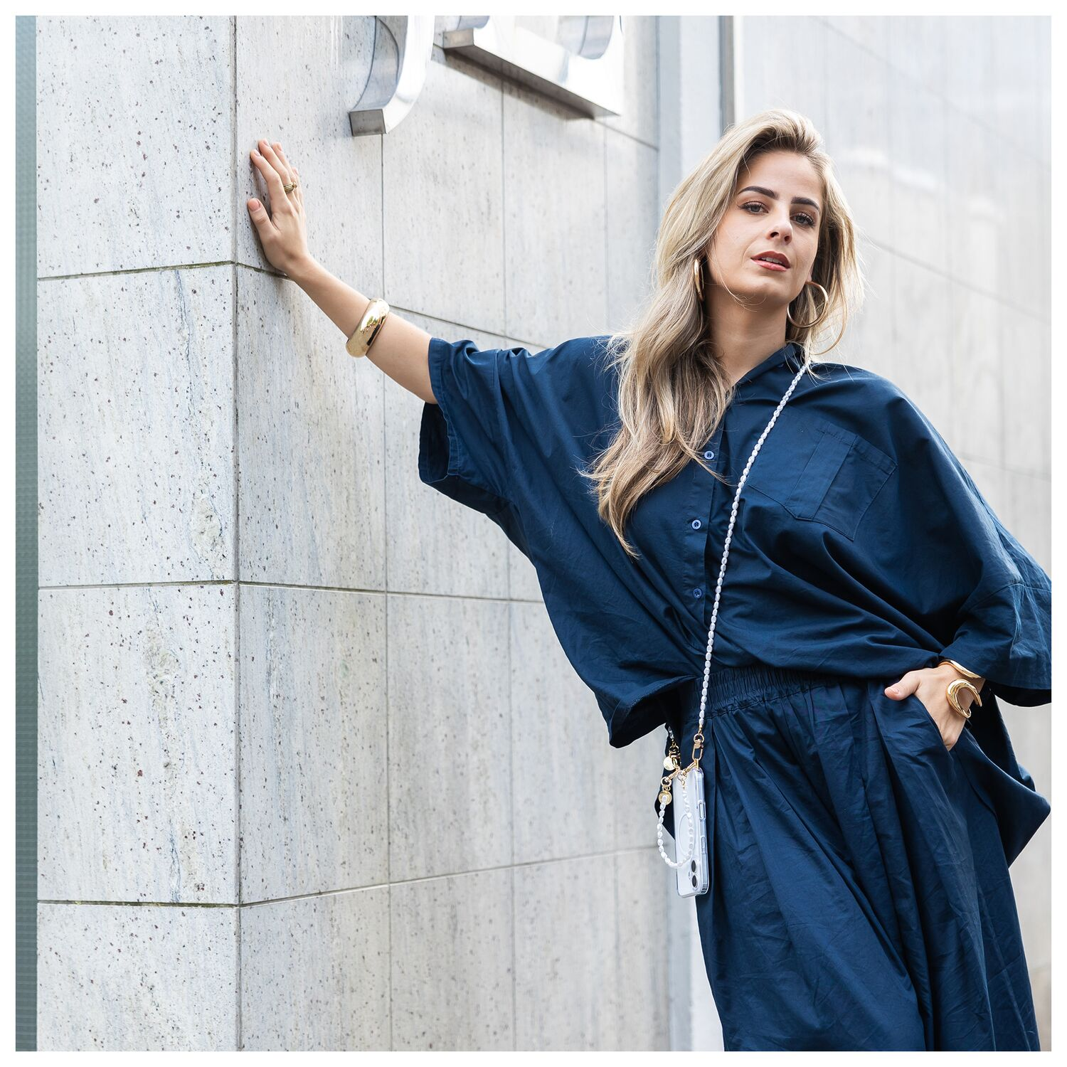 Een blonde vrouw in een donkerblauwe jurk leunt tegen een muur.