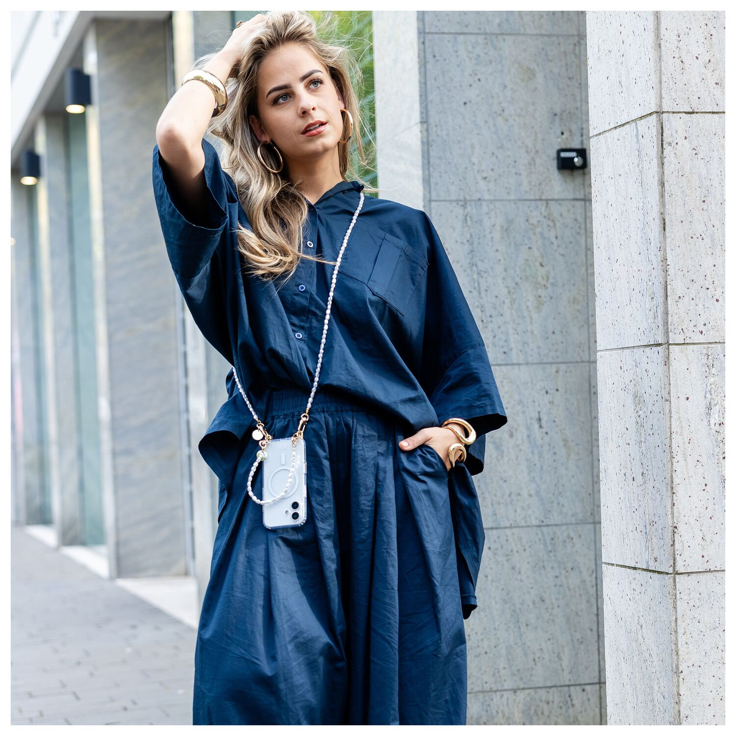 Vrouw in donkerblauw outfit met telefoonketting en sieraden voor stenen gebouw.