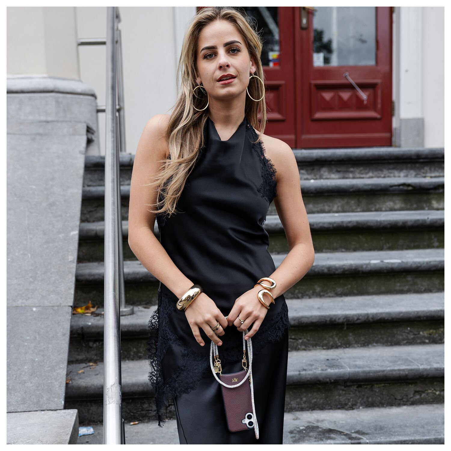 Vrouw in zwarte jurk met gouden sieraden houdt telefoonhoes op trap.