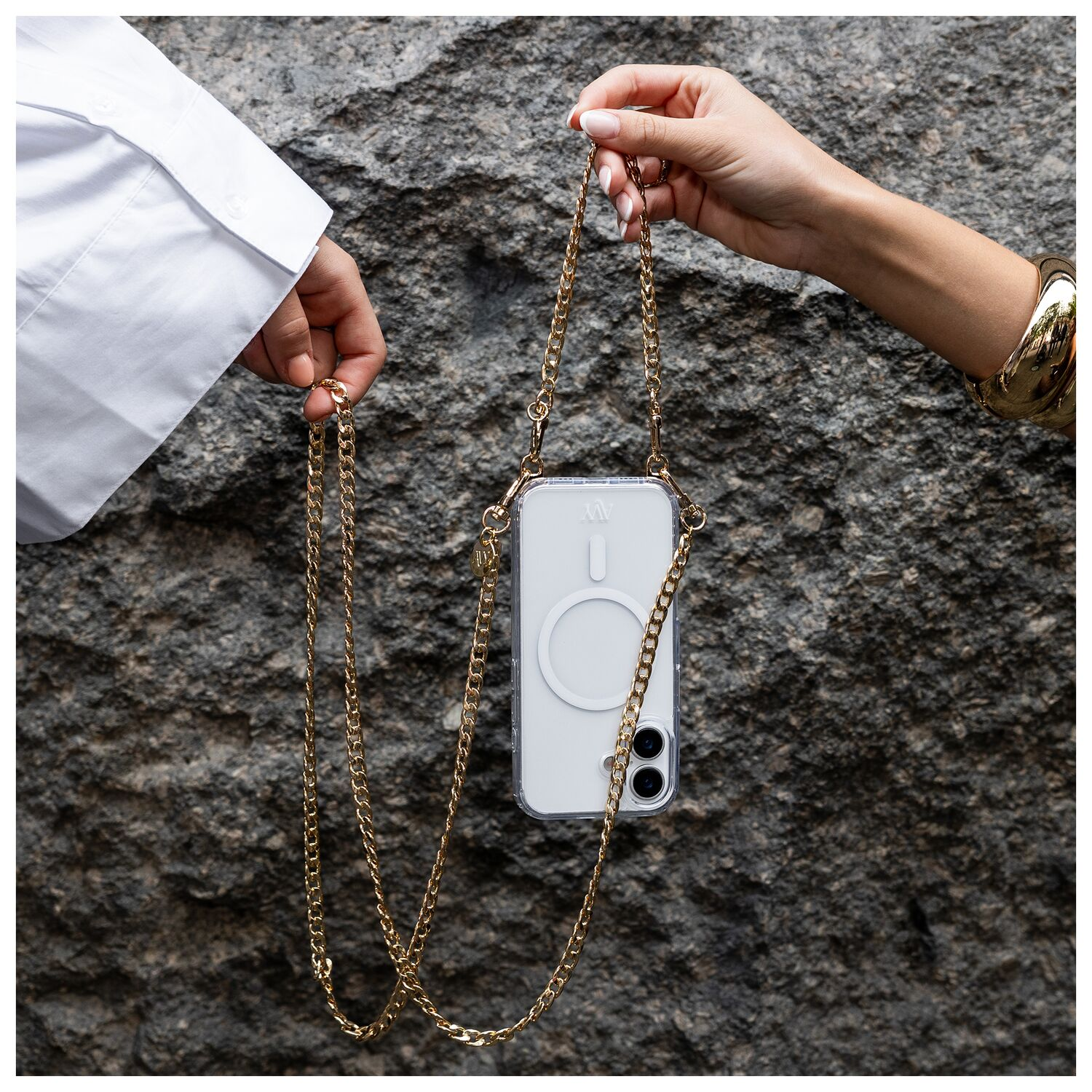 Twee handen houden een doorzichtige telefoonhoes met gouden ketting vast tegen een rotsachtige achtergrond.