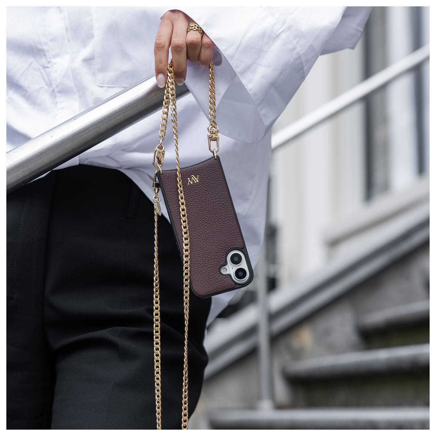 Persoon houdt bruine telefoon met gouden ketting vast. Wit shirt, zwarte broek.