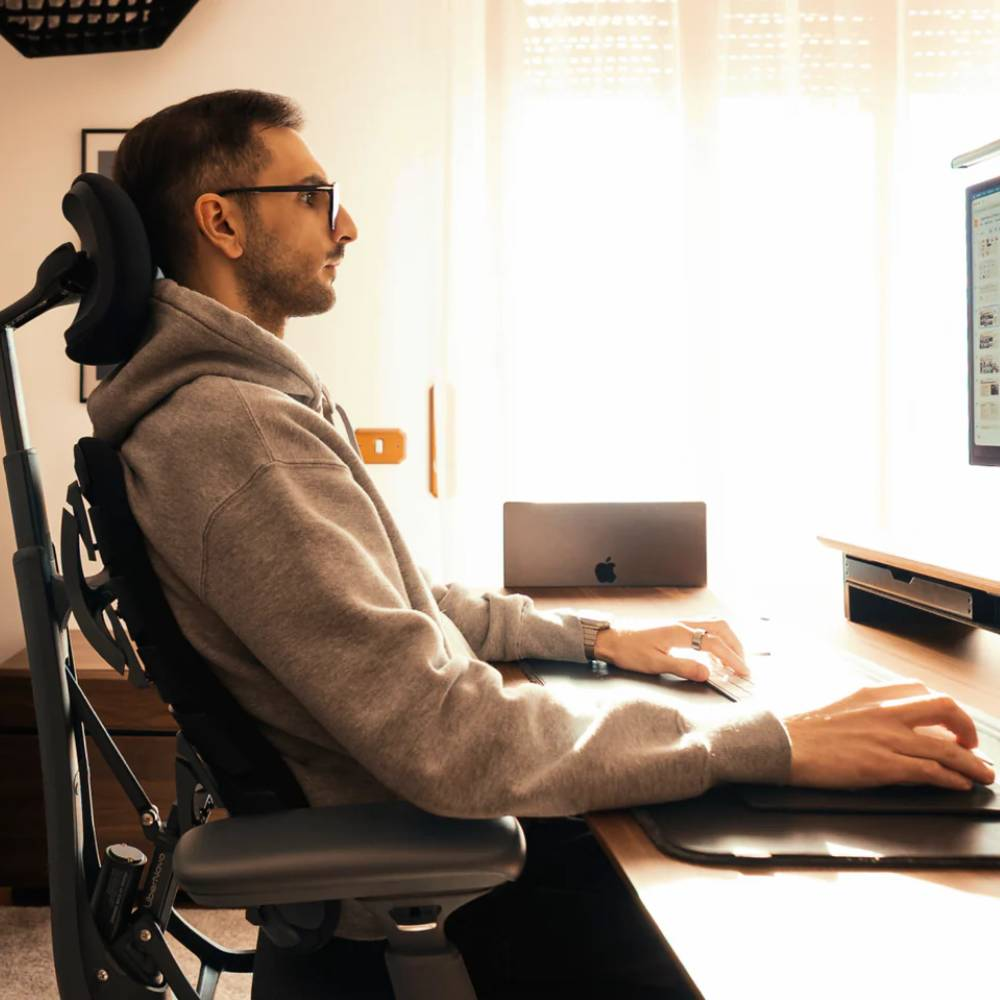 Mann im Kapuzenpullover sitzt an einem Schreibtisch und benutzt eine Maus. Ergonomischer Stuhl.
