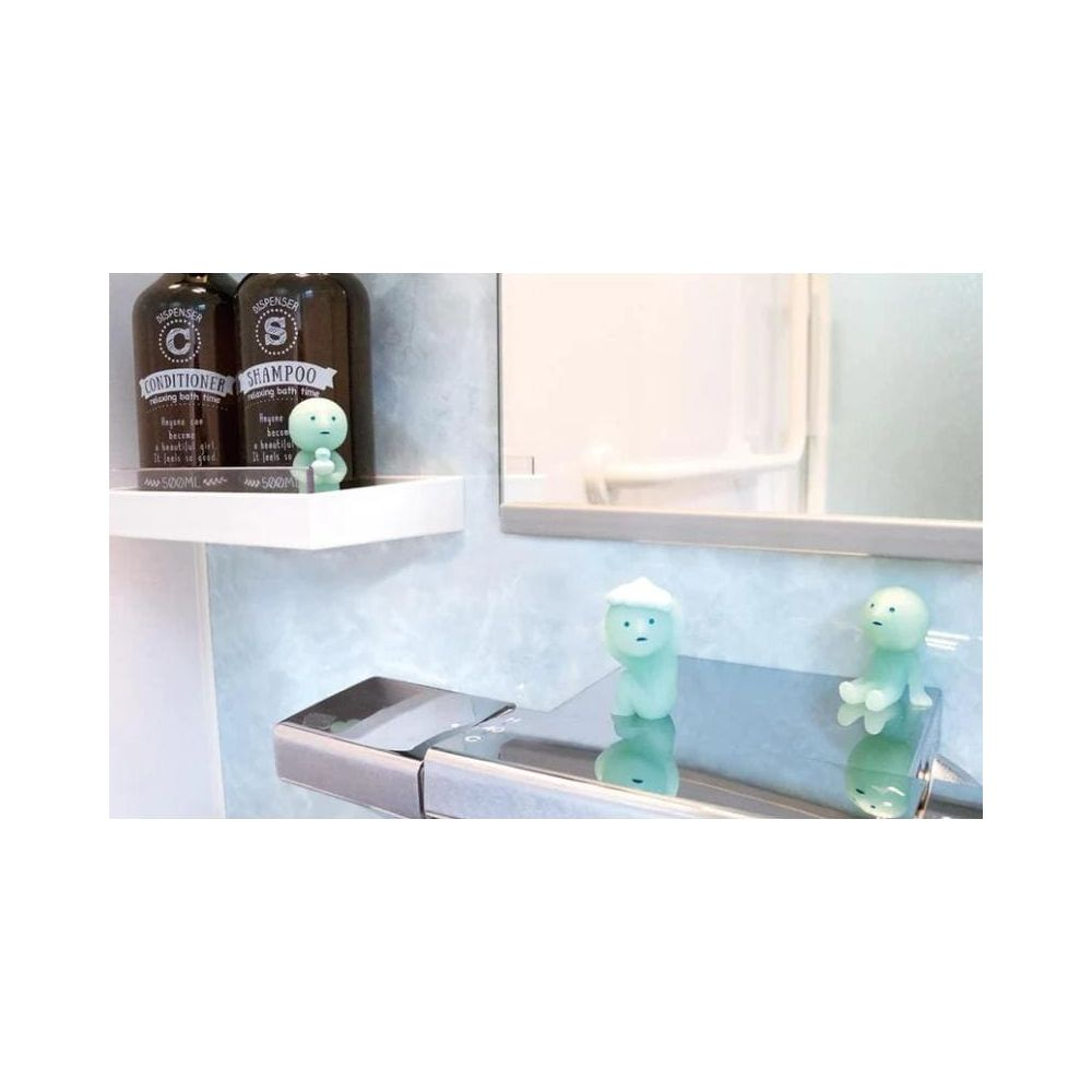 Zwei kleine grüne Figuren stehen auf einem Chromhahn; daneben Shampoo- und Conditioner-Flaschen.