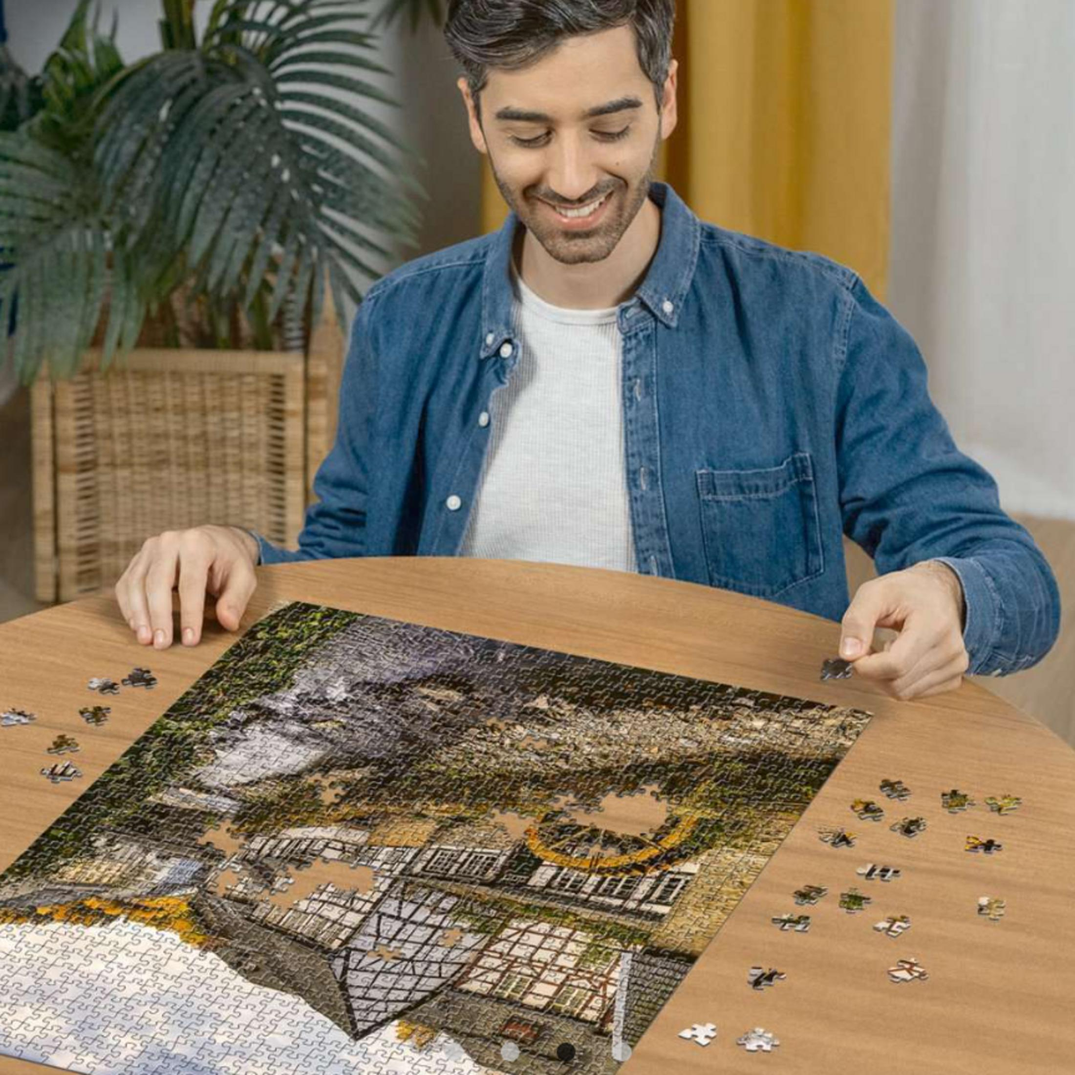 Hombre con camisa vaquera resolviendo un puzzle de una casa y naturaleza. Piezas sueltas en la mesa.