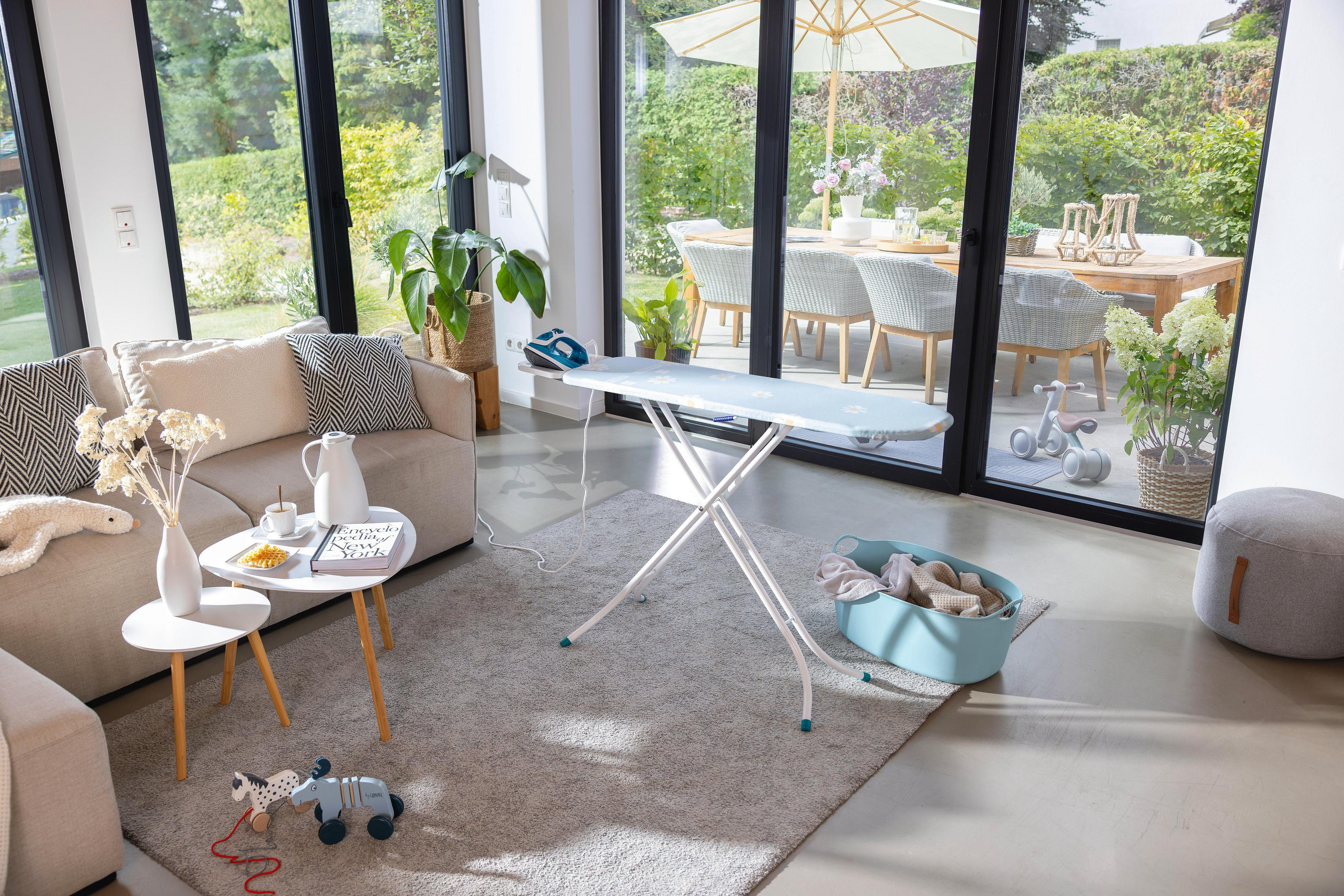 Ein Bügelbrett mit Bügeleisen steht im Wohnzimmer neben einem Sofa und einem Couchtisch.