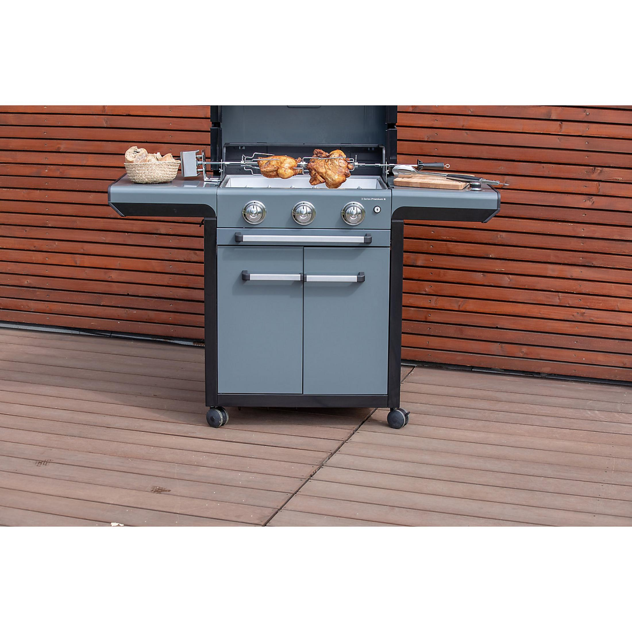 Ein Gasgrill mit zwei Hähnchen am Drehspieß vor einer Holzverkleidung.