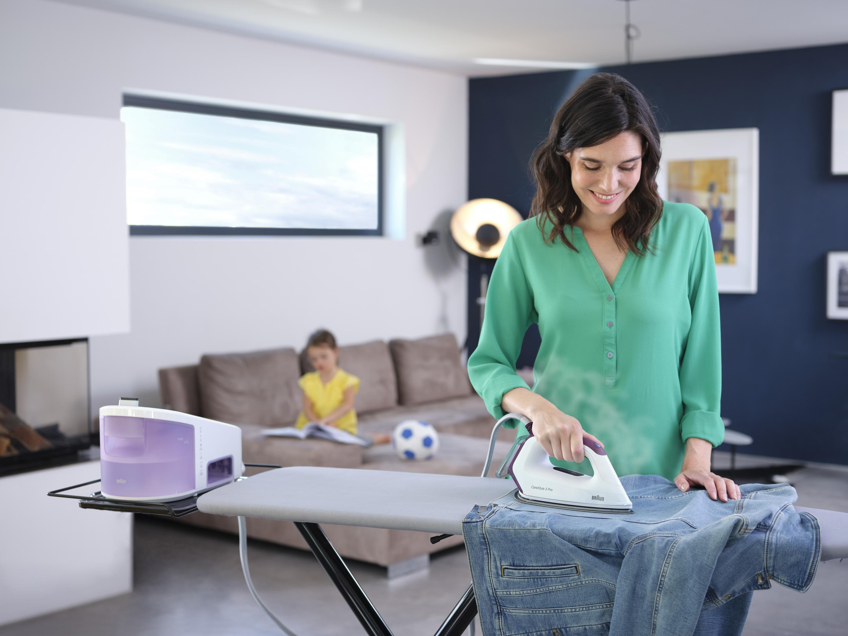 Una donna stira una giacca di jeans su un asse da stiro mentre un bambino legge sullo sfondo.