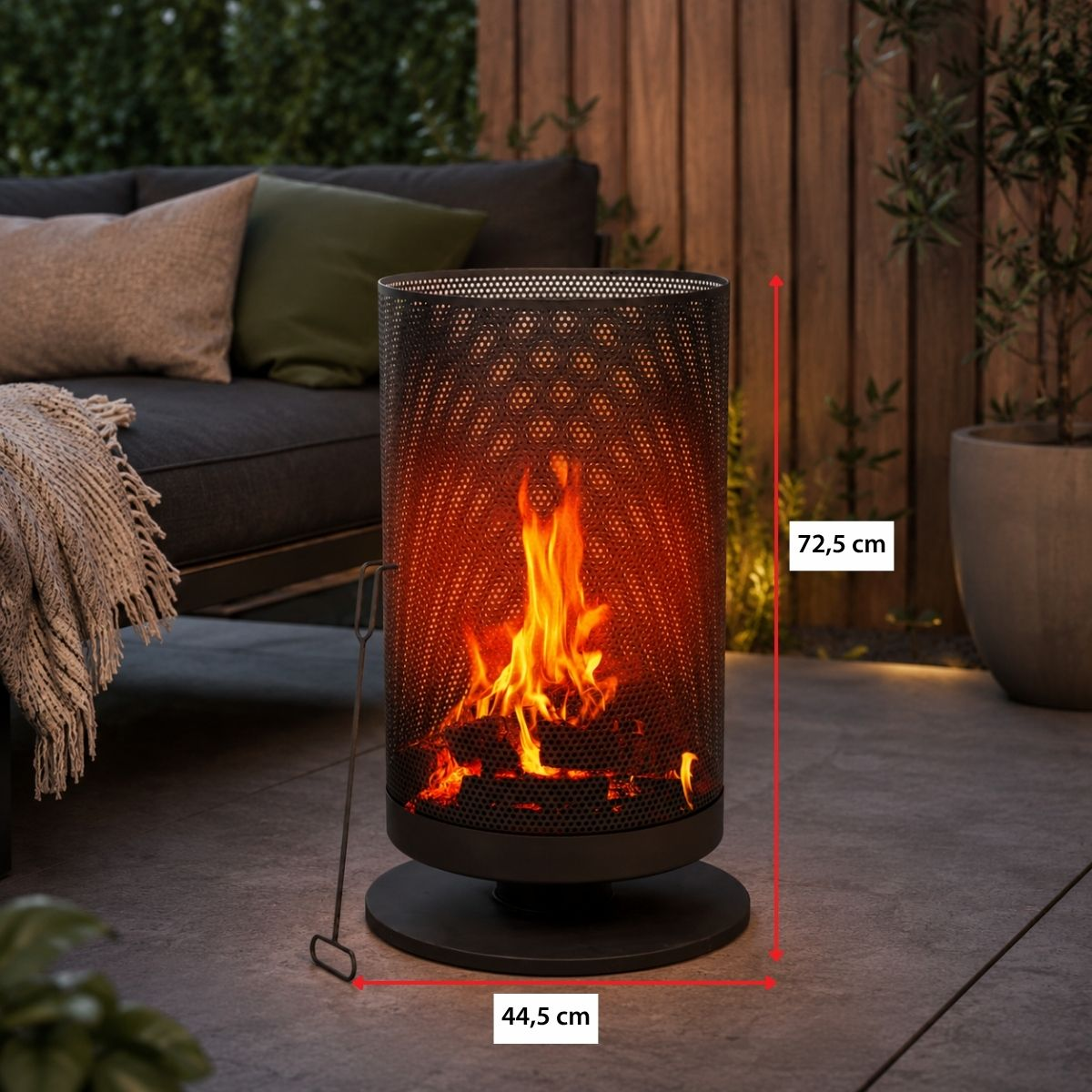 A cylindrical metal fire pit with flames, next to a sofa with pillows and blanket.
