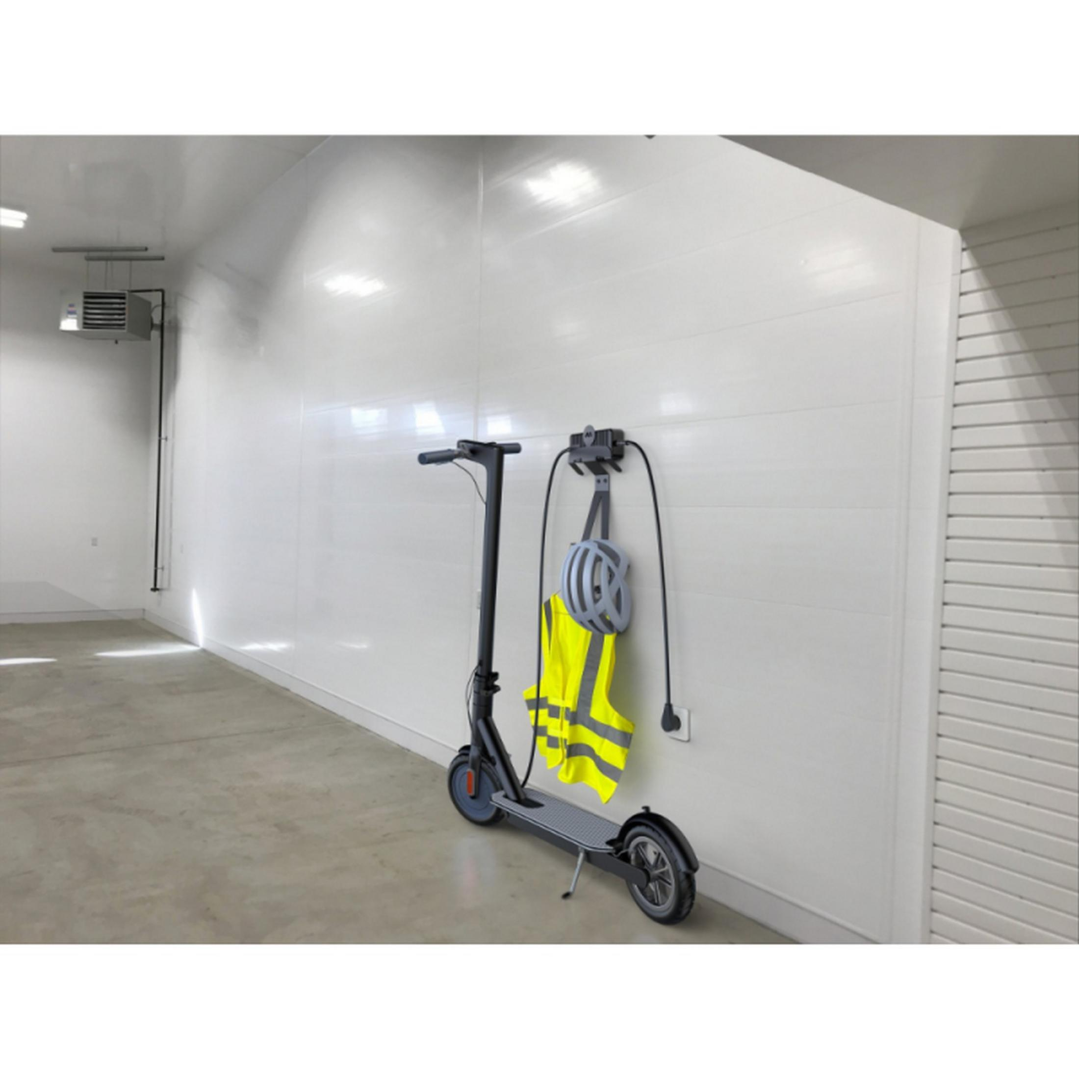Grauer Elektroroller, drinnen geparkt neben einer Wand mit Ladeausrüstung und Warnweste.