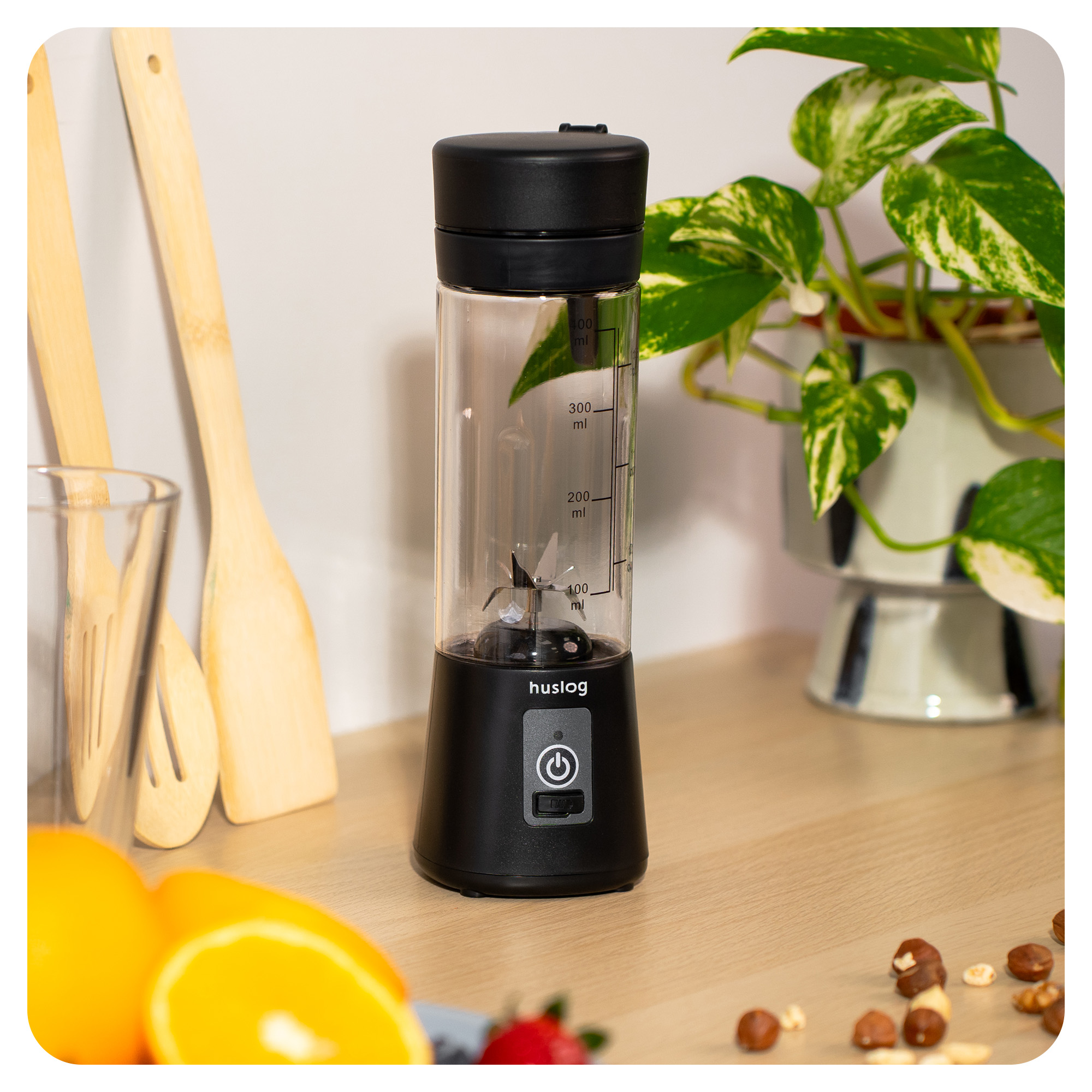 Przenośny blender z czarnym i przezroczystym pojemnikiem, obok pomarańczy i orzechów.