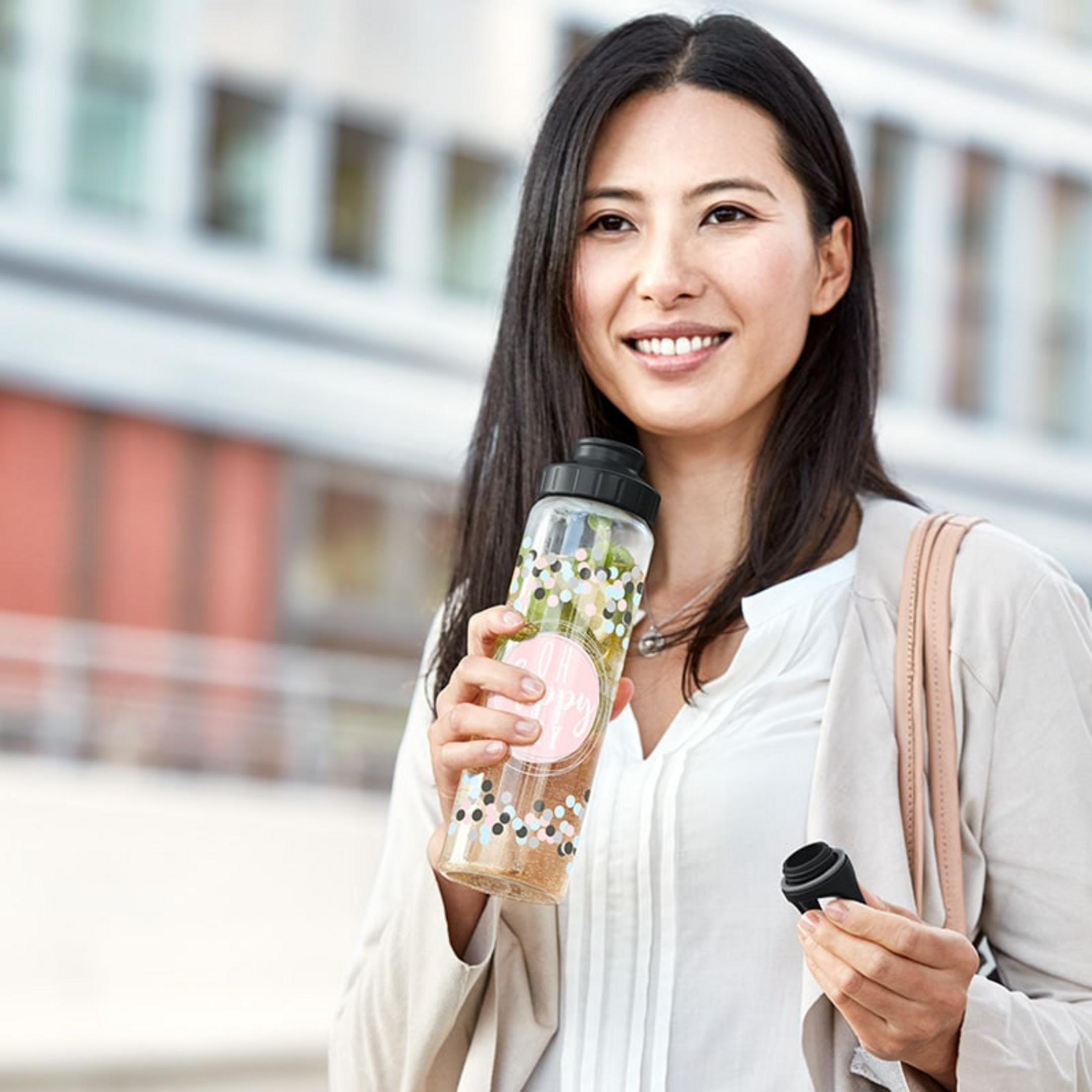 Femme tenant une gourde et un sac, souriant devant un bâtiment.