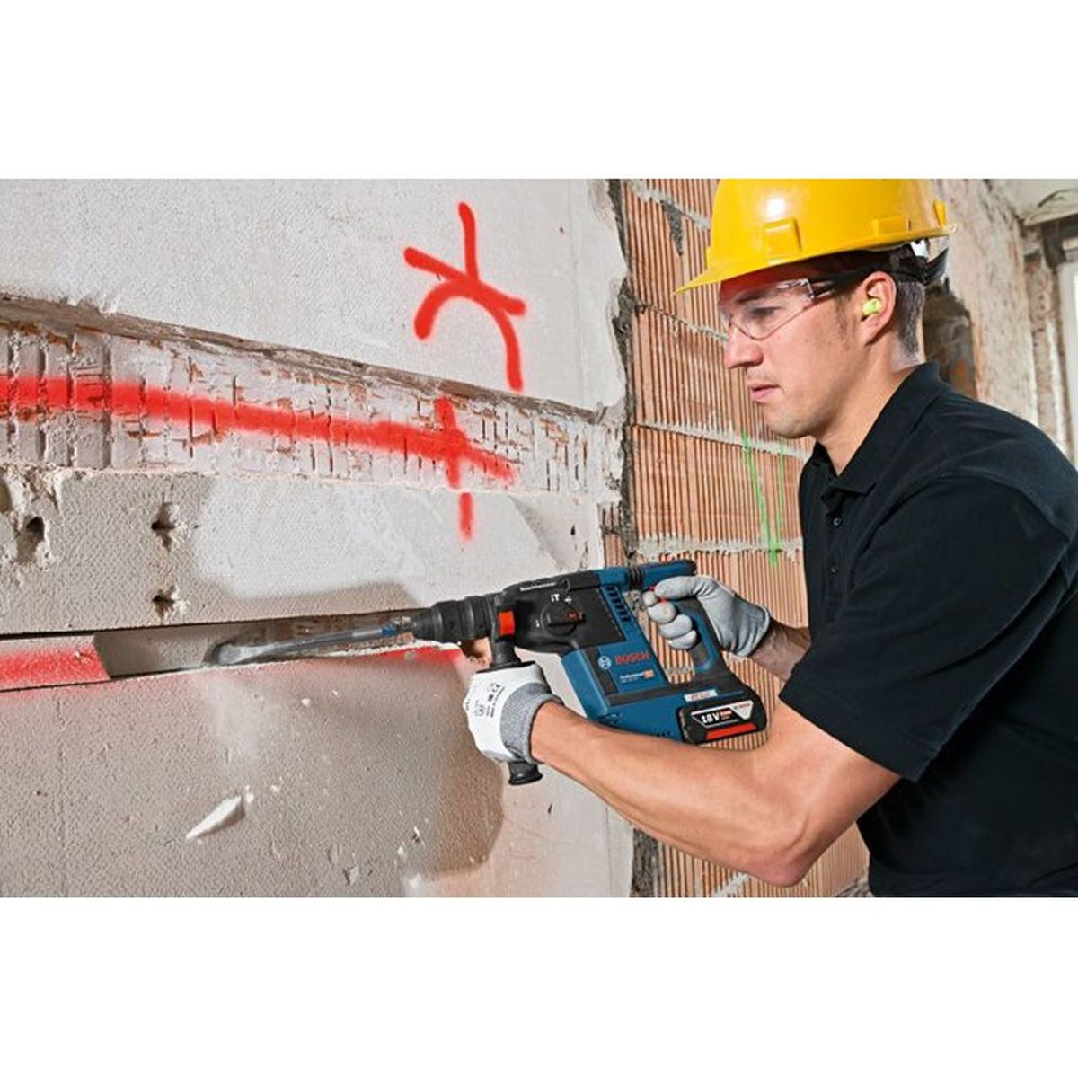 Mann mit Bohrhammer arbeitet an Wand mit roter Markierung, Schutzbrille und Helm.