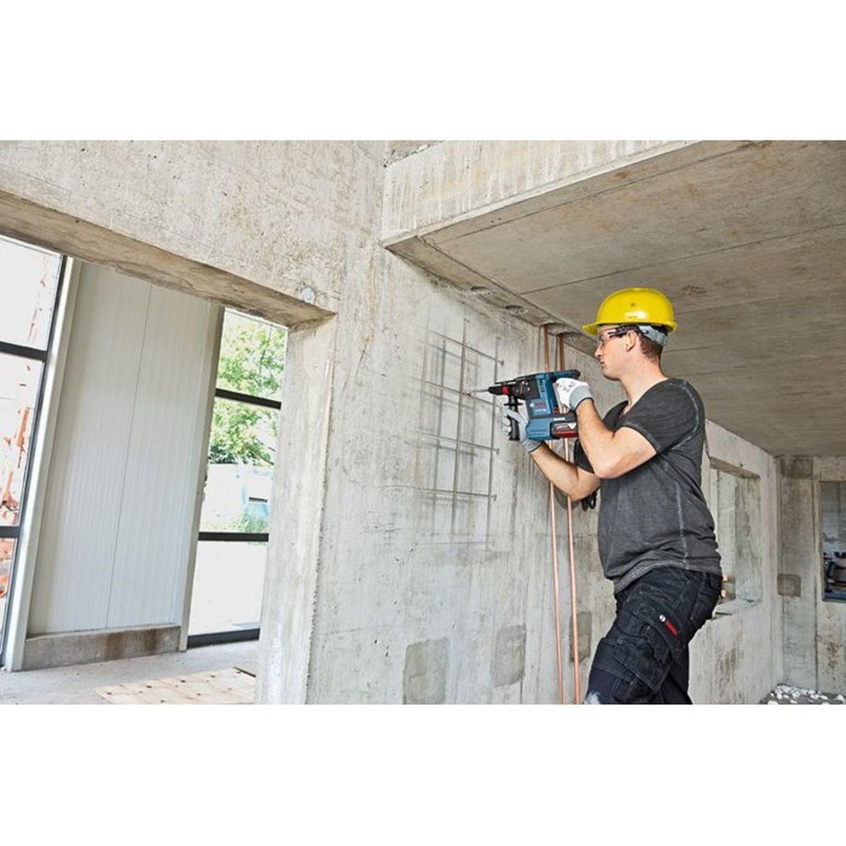 Ein Bauarbeiter mit gelbem Helm bohrt mit einem blauen Bohrer in eine Betonwand.