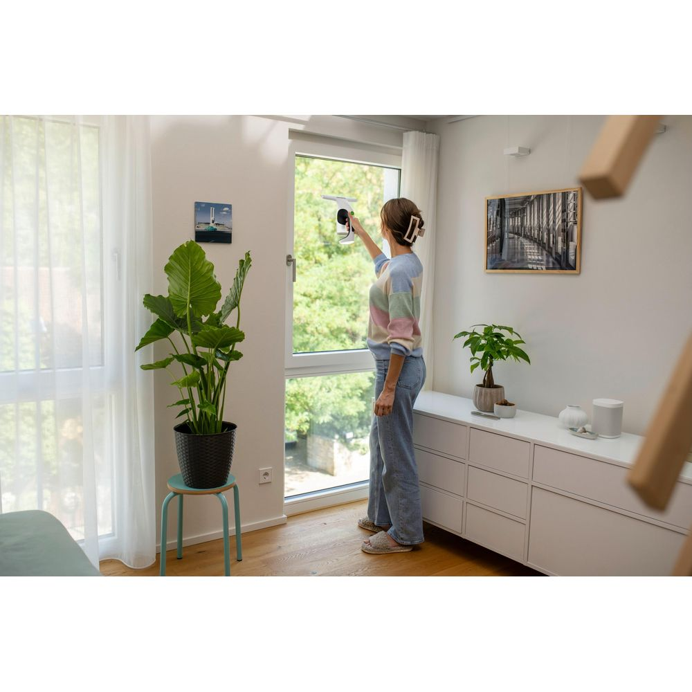 Frau reinigt Fenster mit einem Akku-Fenstersauger. Sie trägt einen gestreiften Pullover und Jeans.