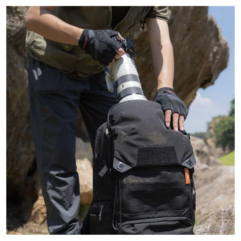 Mann in Handschuhen packt Objektiv in Tarnrucksack vor Felsen.