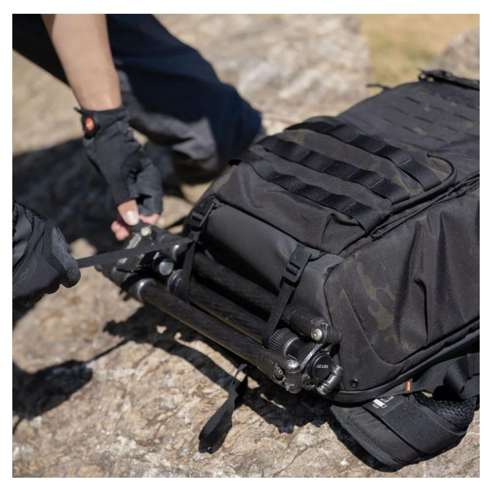Person befestigt Stativ am schwarzen Rucksack mit Tarnmuster auf felsigem Untergrund.