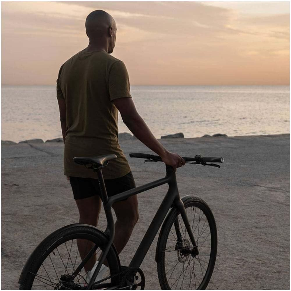 Mann in olivgrünem Hemd und schwarzen Shorts hält ein schwarzes Fahrrad am Meer bei Sonnenuntergang.