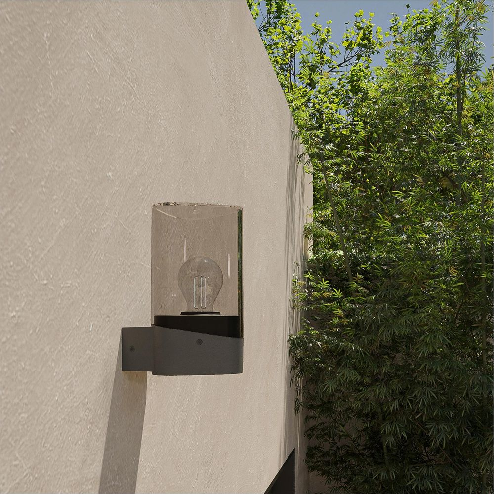 Eine Wandleuchte mit Glühbirne und Glaszylinder an einer cremefarbenen Wand neben grünen Bäumen.