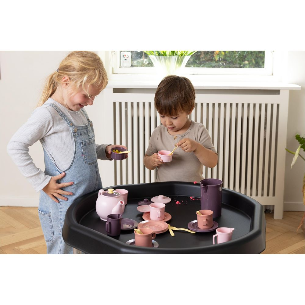 Zwei Kinder spielen mit einem Spielzeug-Teeservice auf einem schwarzen Tisch. Sie halten kleine Löffel und Tassen.