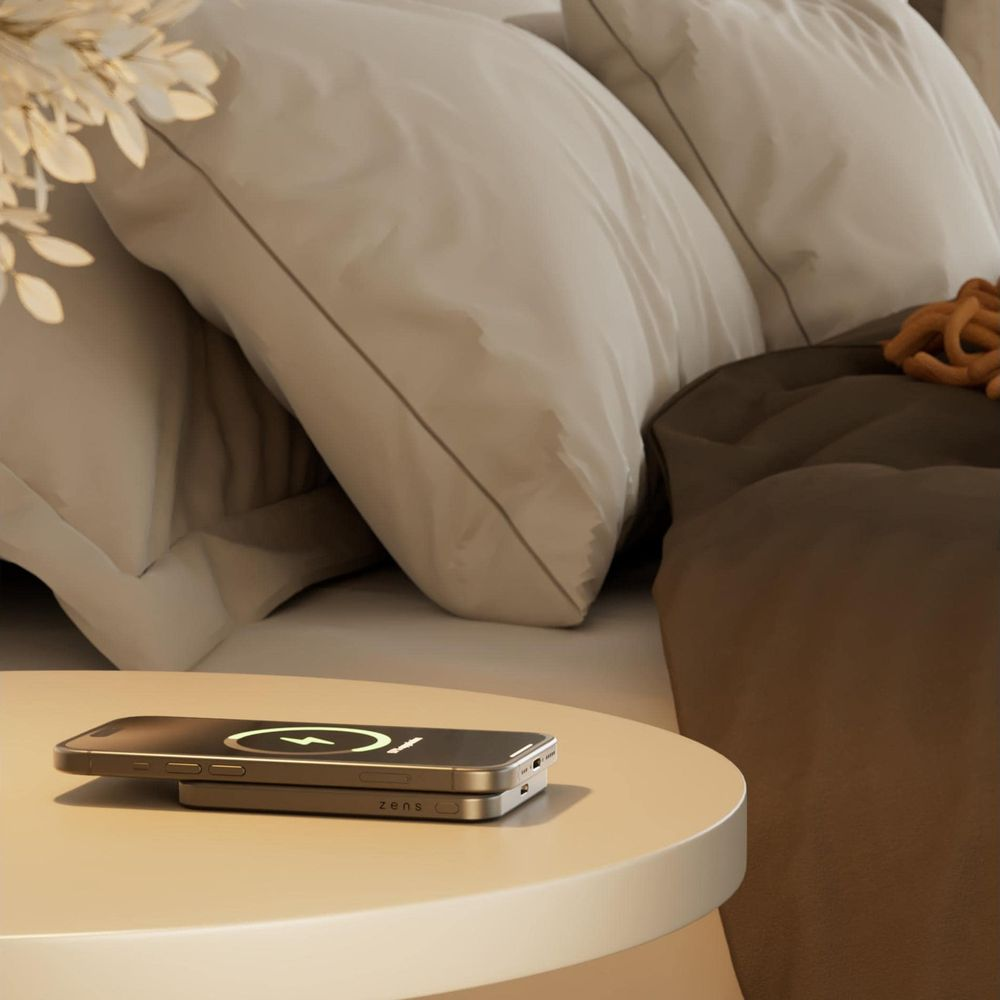 Ein Smartphone und ein Ladegerät liegen auf einem Beistelltisch neben einem Bett.