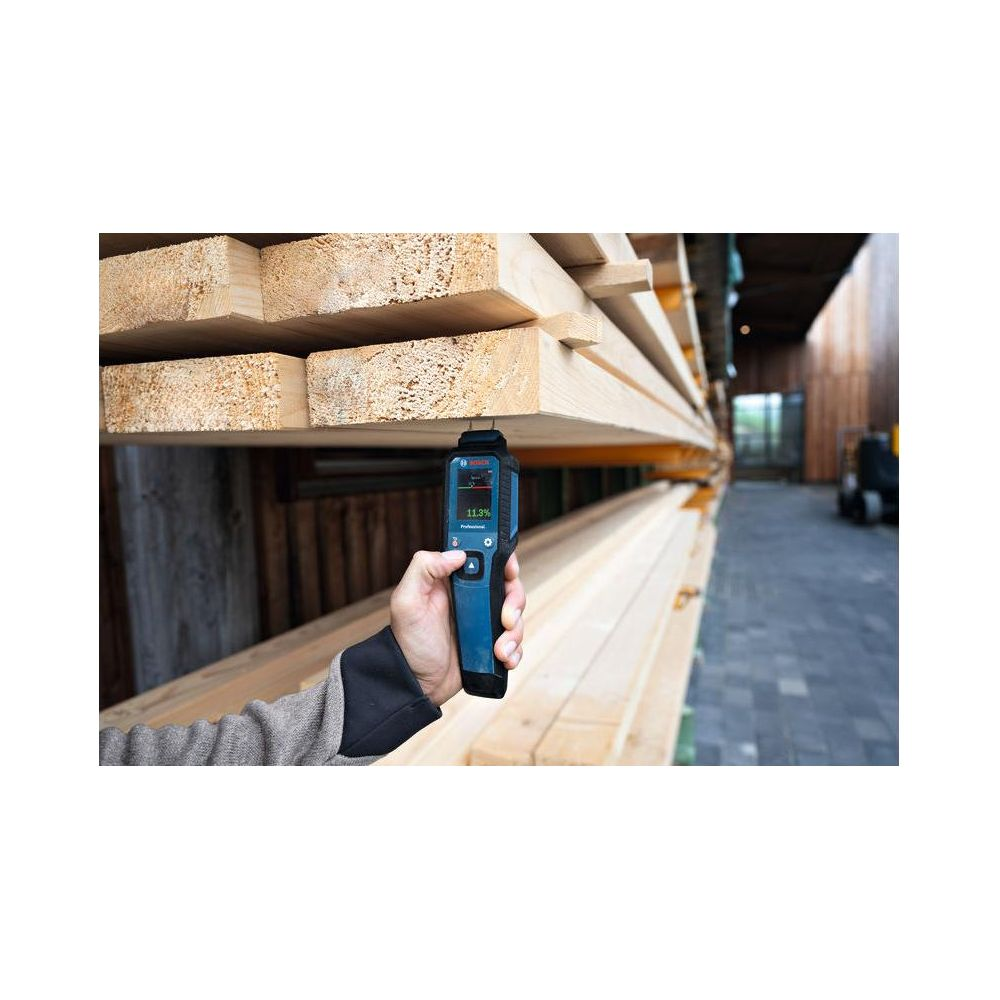 Ein Messgerät prüft Holz auf Feuchtigkeit. Holzstapel im Hintergrund.