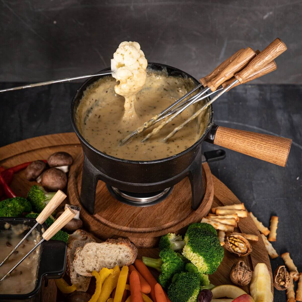 Käsefondue mit Brot, Gemüse, Pilzen und Nüssen auf einem Holzteller.