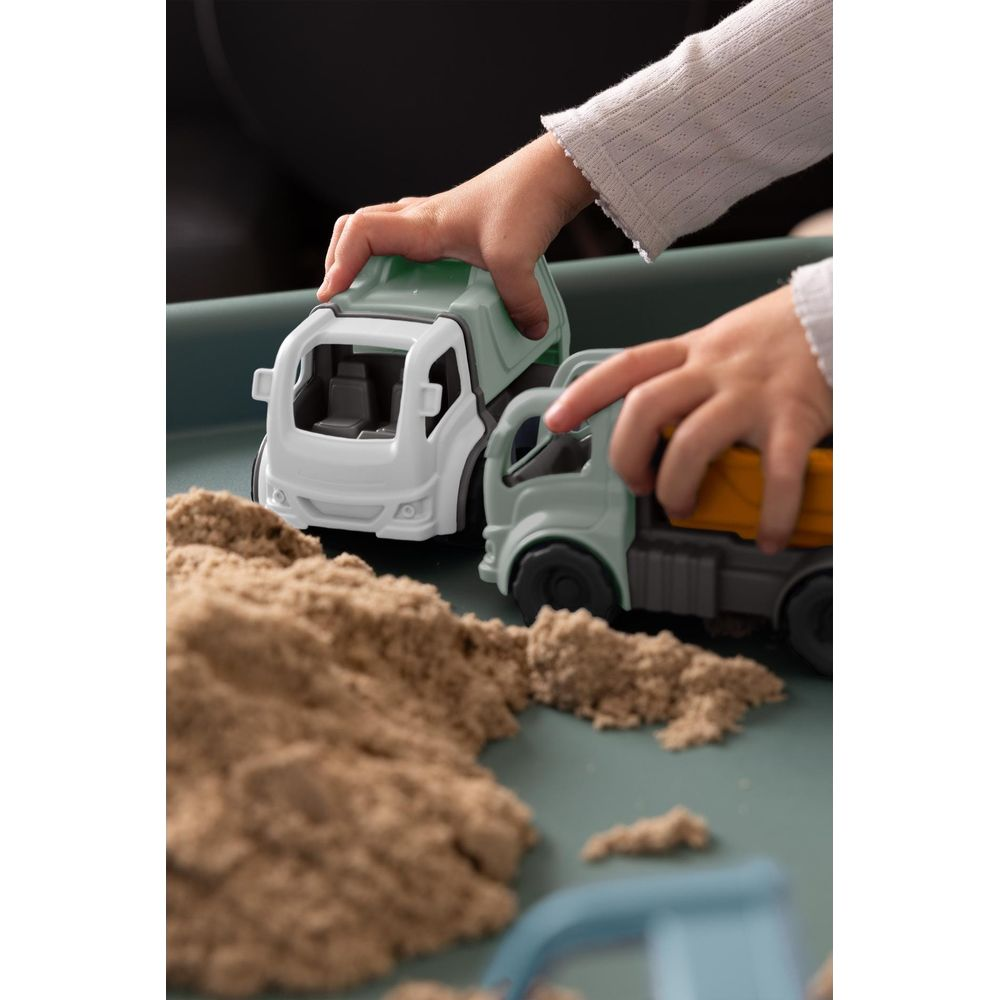 Kinderhände spielen mit hellgrünen und weißen Spielzeuglastwagen im Sand.
