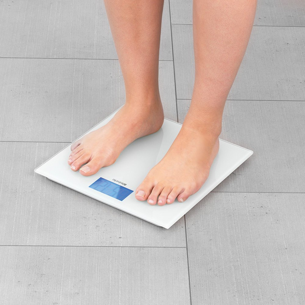 Nackte Füße auf einer weißen Digitalwaage, zeigt 67,5. Grauer Fliesenboden.