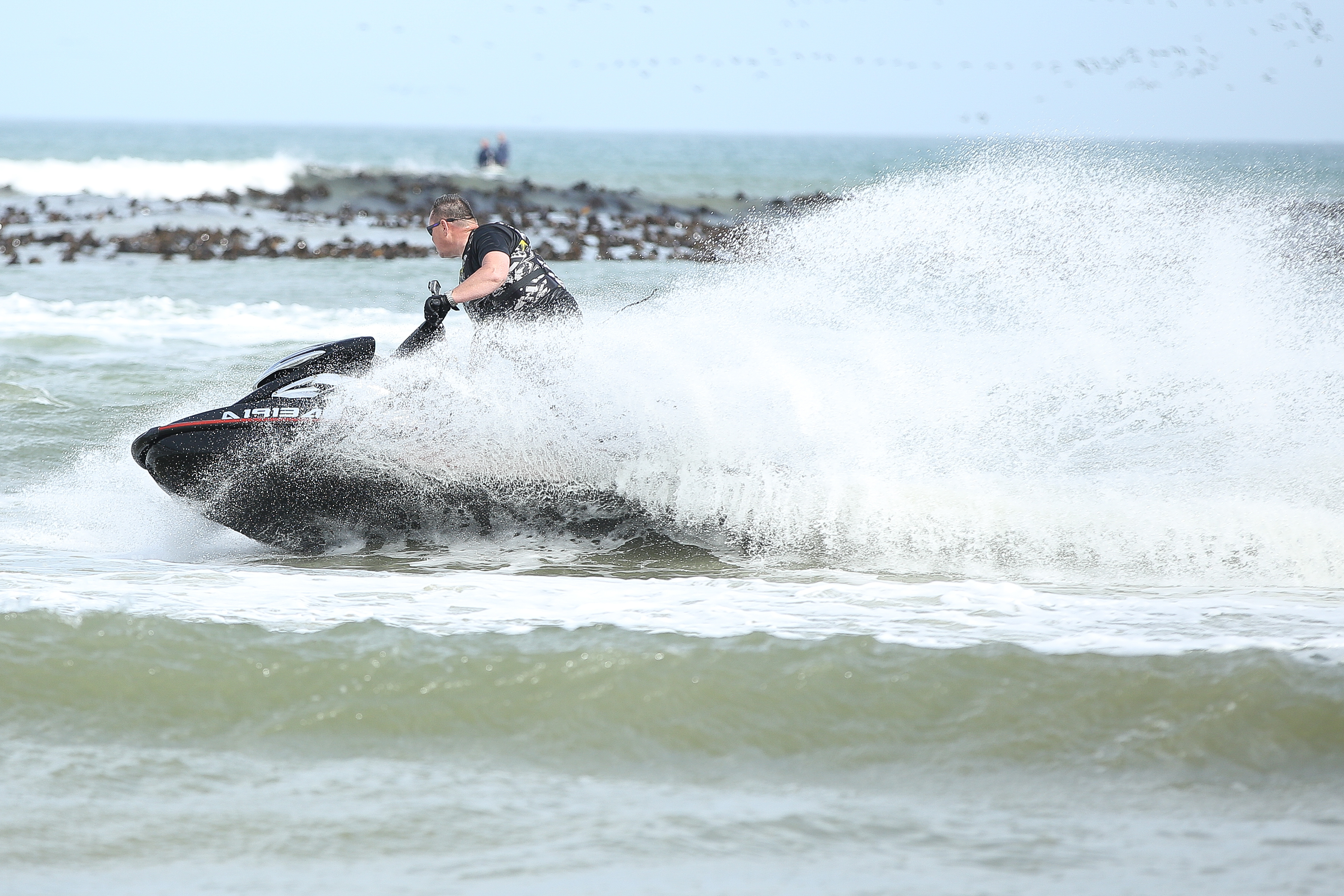 Mann fährt Jet-Ski mit viel Gischt, im Hintergrund Felsen und Meer.