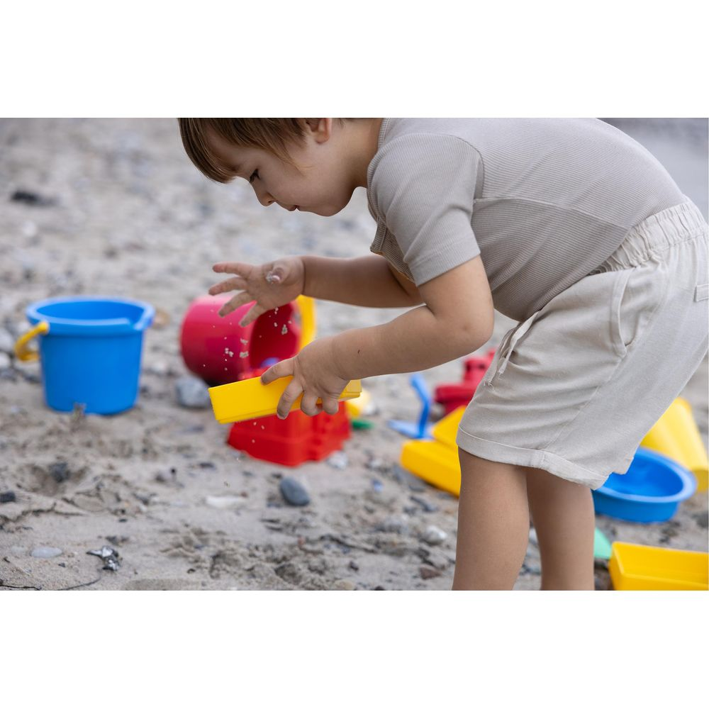 Ein Kind spielt mit bunten Sandspielzeugen am Strand. Ein blauer Eimer steht links.