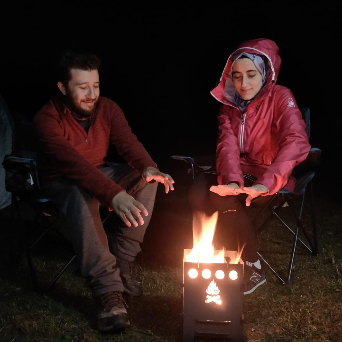 İki kişi gece kamp ateşinin yanında oturuyor, ellerini ısıtıyor.