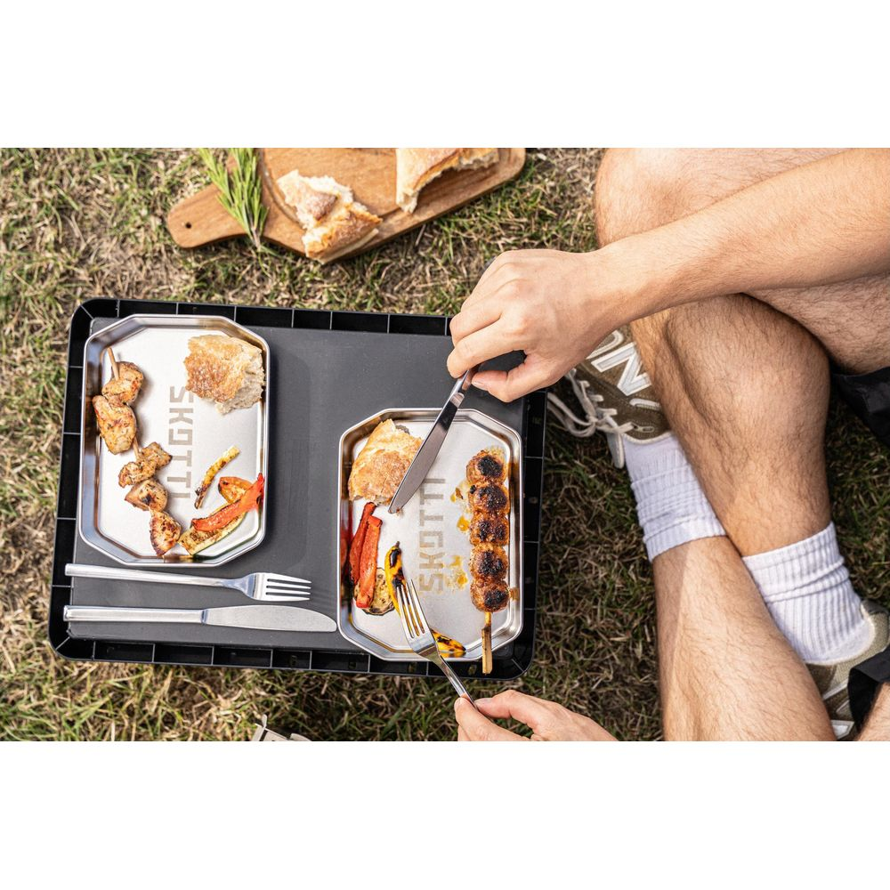 Zwei Personen essen Gegrilltes mit Brot und Gemüse auf einem Tablett im Freien.