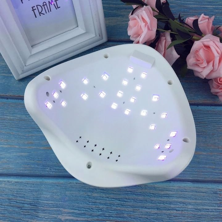 Eine weiße UV-Nagellampe mit LEDs auf einer blauen Holzoberfläche, neben rosa Rosen und einem Rahmen.