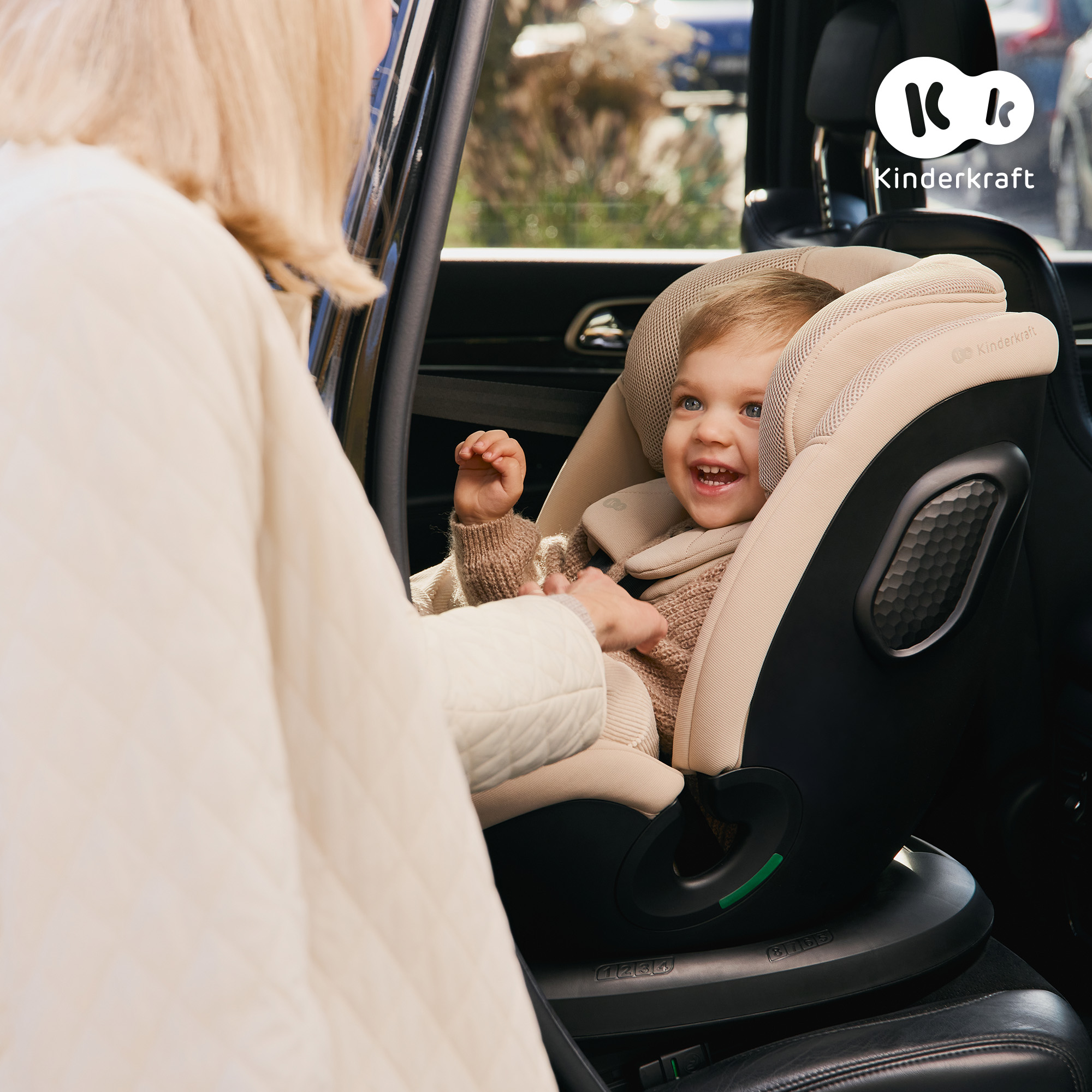 Frau schnallt Kind in beigen Autositz. Kind lächelt. Kinderkraft Logo.