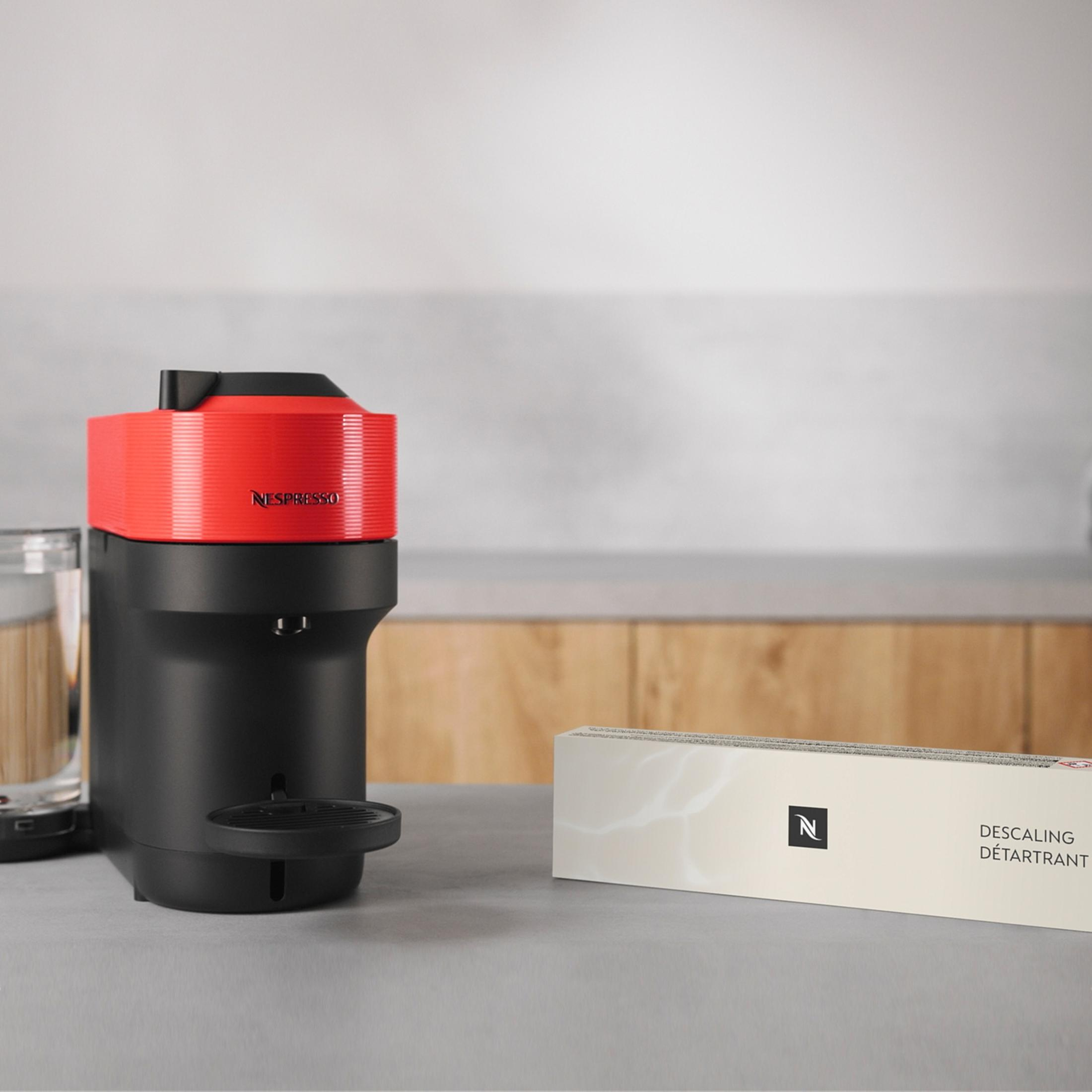 Una macchina Nespresso rossa e nera con un bicchiere e una scatola decalcificante accanto.