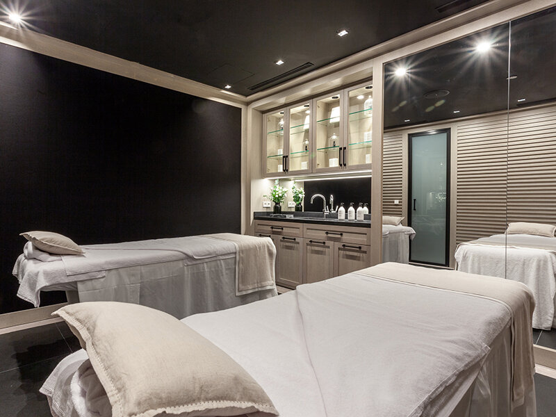 Dos camillas de masaje blancas están dispuestas en una sala de spa con un estante negro y área de lavabo.