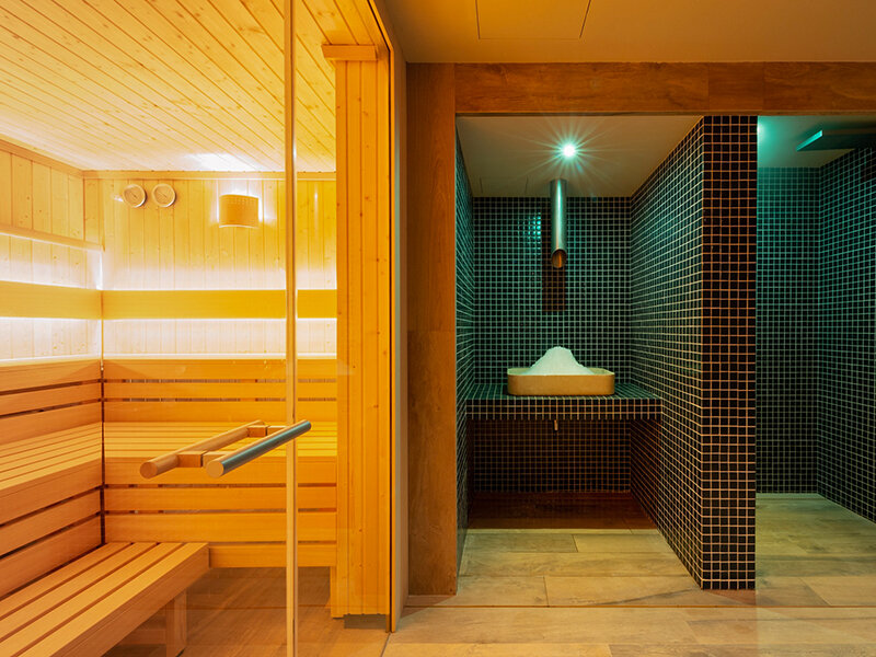 Sauna de madera junto a una sala alicatada con cuenco de sal y ducha.
