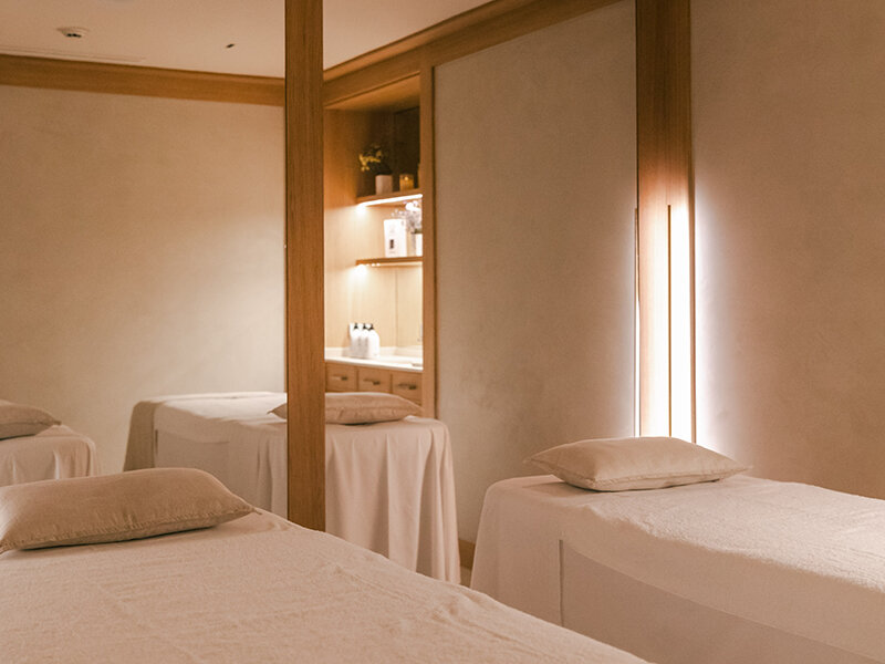 Una habitación luminosa con tres camillas de masaje blancas, cojines e iluminación indirecta.